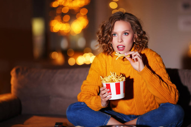 Fascinada joven hembra en suéter comiendo papas fritas y viendo interesante película en la televisión mientras está sentada en el sofá por la noche en hom - Foto, imagen