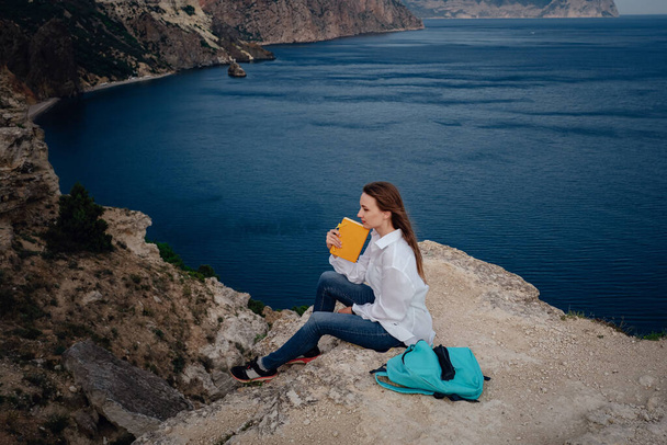Attractive woman is sitting on the edge of the mountain with a sea view, reading a book. Self-education in the open air. writing notes in diary, - Photo, Image