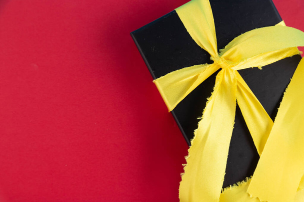 Black gift box with gold satin ribbon on red background. - 写真・画像