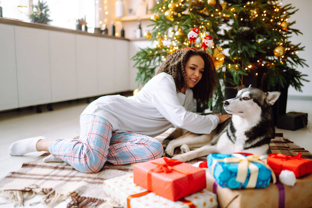 Beautiful woman playing and having fun with her dog while sitting near the christmas tree. Young lady hugging her dog with red christmas hat. Winter holidays. Light around. - Foto, immagini