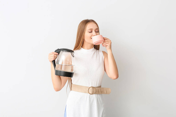Ευτυχισμένη γυναίκα κρατώντας ηλεκτρικό βραστήρα και πίνοντας τσάι σε λευκό φόντο - Φωτογραφία, εικόνα
