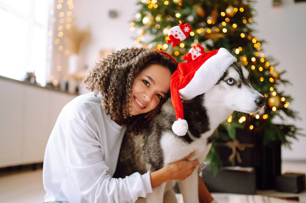 Beautiful woman playing and having fun with her dog while sitting near the christmas tree. Young lady hugging her dog with red christmas hat. Winter holidays. Light around. - Foto, Imagem