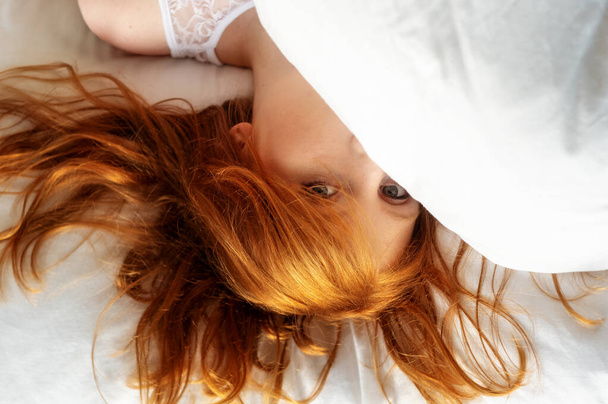 View from above to an attractive, young, sexy redhead, red-haired woman, hair wild on the sheets, face half under the pillow, lying in fresh soft white sheets in the bedroom, copy space. - Foto, immagini