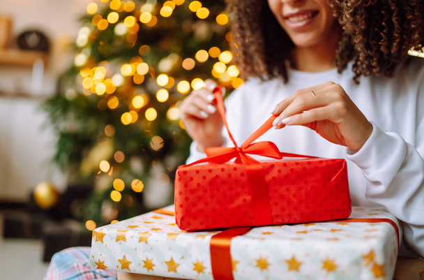 Χριστούγεννα ή Πρωτοχρονιά κουτί δώρου στα γυναικεία χέρια. Η ευτυχισμένη γυναίκα ανοίγει δώρα κοντά στο χριστουγεννιάτικο δέντρο. Χειμερινές διακοπές. - Φωτογραφία, εικόνα