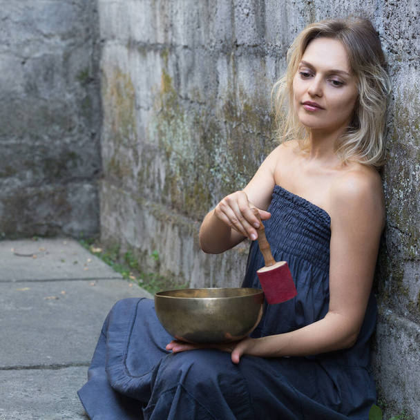 Beautiful happy young woman is sitting and using a singing bowl. Medetation yoga concept. Grey blue colors. - Фото, изображение