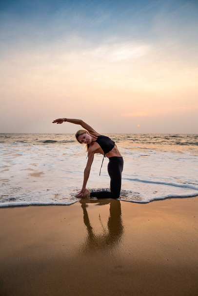 Mulher de meia idade em preto fazendo ioga na praia de areia na Índia horário do pôr do sol. - Foto, Imagem