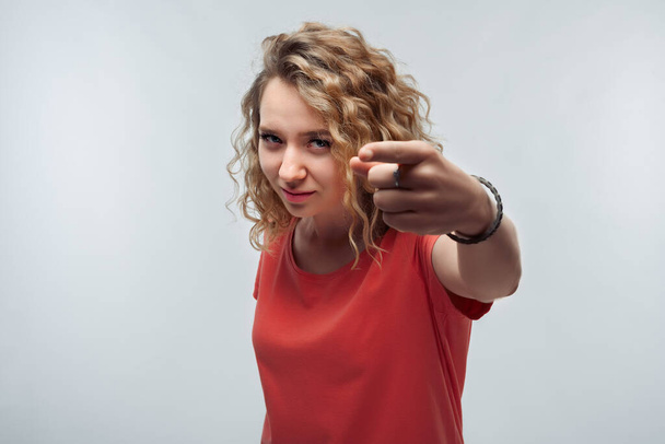 Pretty Young woman with curly hair in casual t shirt looking at the camera and pointing her finger at you. Studio shot, white background. Human emotions, facial expression concept - Fotoğraf, Görsel