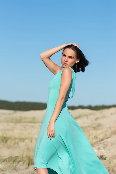 Mujer sensual delgada de cuerpo completo con pelo volador que usa vestido largo de turquesa mientras está de pie en el viento en la costa - Foto, imagen