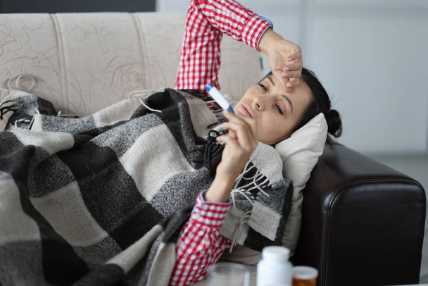 Erkältete Frau liegt auf Sofa unter Decken und schaut auf Thermometer - Foto, Bild