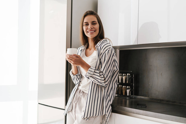 Young beautiful caucasian woman smiling and drinking coffee at home - 写真・画像