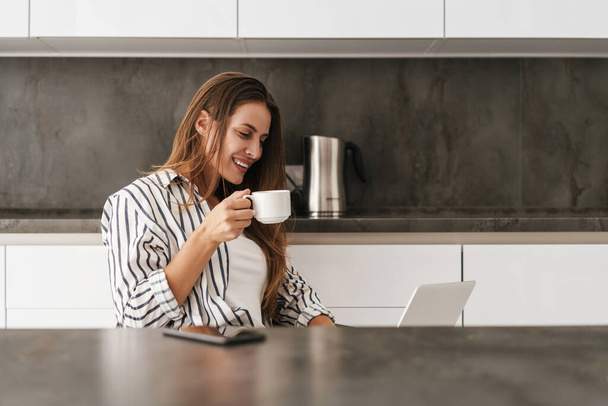 Young beautiful caucasian woman working on laptop and drinking coffee at home - Foto, imagen