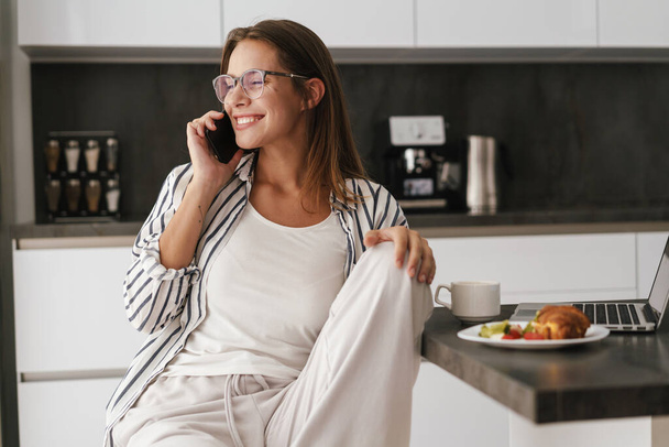 Young beautiful caucasian woman talking on cellphone while having breakfast at home - Photo, Image