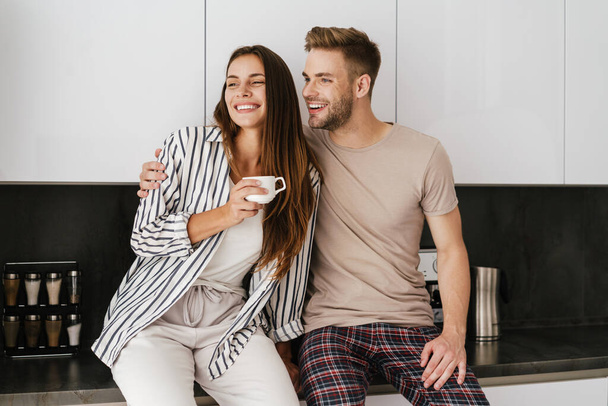 Young beautiful caucasian couple in leisure clothes smiling and drinking coffee at home - 写真・画像