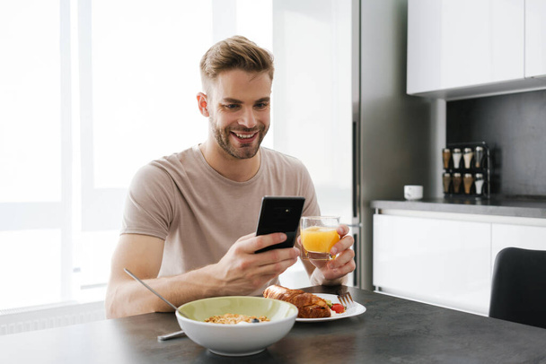 Young handsome caucasian man holding cellphone while having breakfast at home - Foto, Imagem
