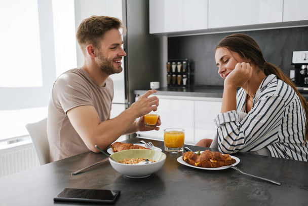 Young beautiful caucasian couple sitting at table while having breakfast at home - 写真・画像