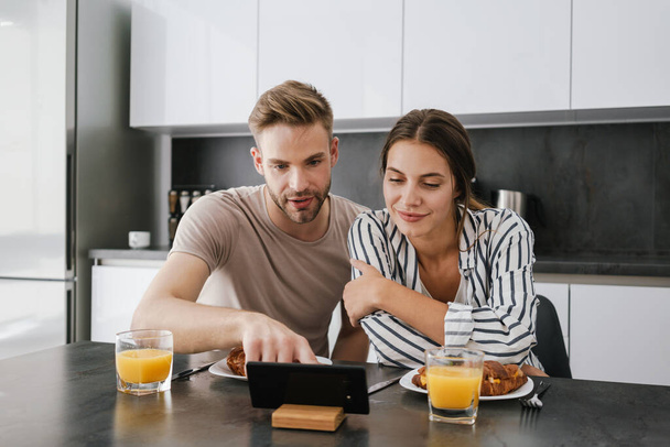 Young caucasian couple looking at cellphone together while having breakfast at home - Photo, image