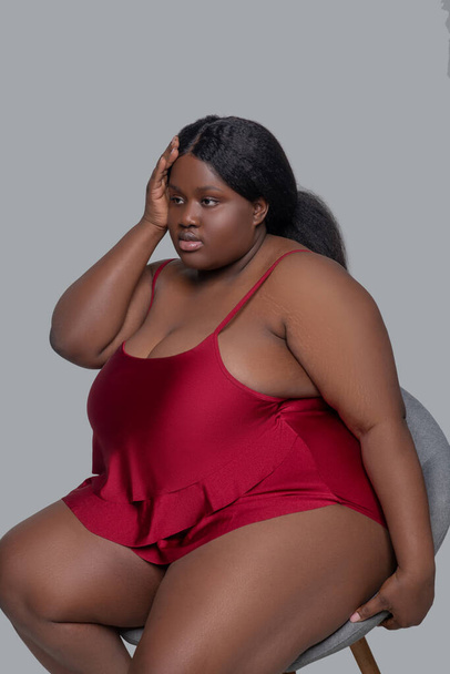 Plus size african american young woman in red underwear sitting on a chair and looking upset - Foto, imagen