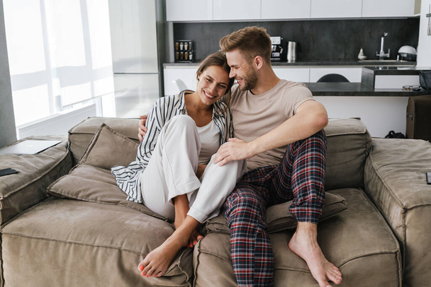 Young beautiful caucasian couple smiling and hugging together while sitting on sofa at home - 写真・画像