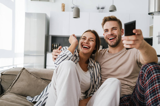 Young caucasian couple sitting on sofa and holding remote control while watching television at home - Foto, Bild