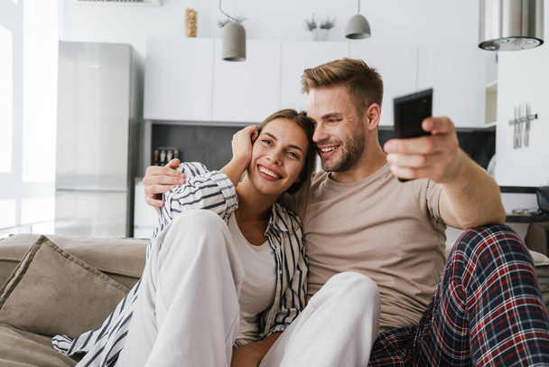 Young caucasian couple sitting on sofa and holding remote control while watching television at home - Foto, Imagem