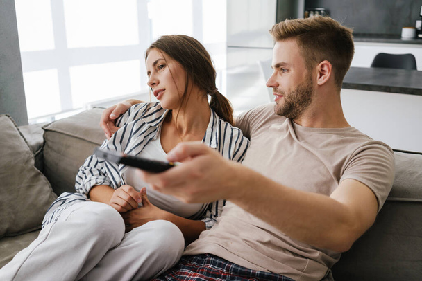 Young caucasian couple sitting on sofa and holding remote control while watching television at home - Fotó, kép