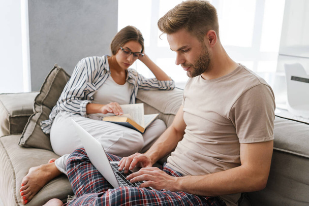Young caucasian couple holding laptop and reading book while sitting on sofa at home - Photo, image
