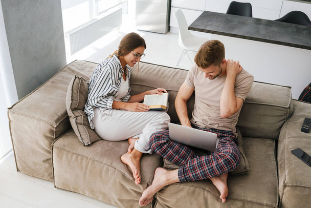 Young caucasian couple holding laptop and reading book while sitting on sofa at home - Photo, Image