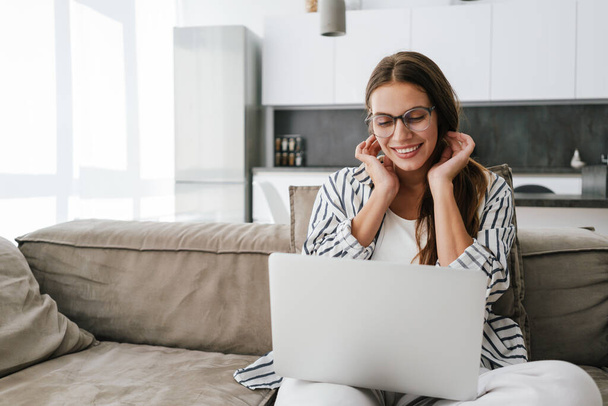 Young caucasian happy woman smiling and using laptop while sitting on sofa at home - Foto, Imagem