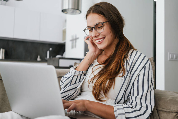 Young caucasian happy woman smiling and using laptop while sitting on sofa at home - Foto, Bild