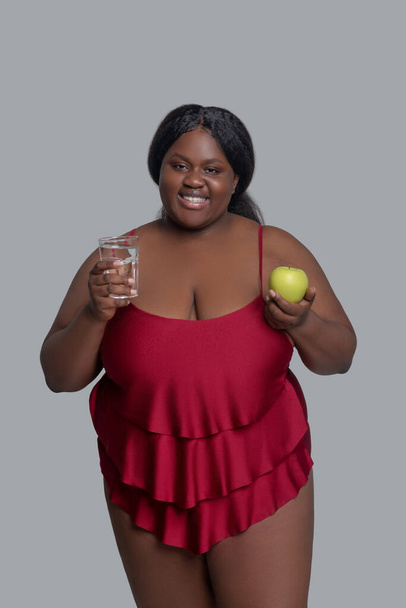 Tummaihoinen hymyilevä nainen, jolla on omena ja lasi vettä. - Valokuva, kuva