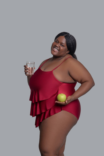 Dark-skinned woman holding an apple and a glass of water and smiling - Фото, изображение