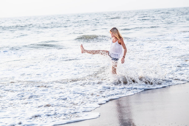 Aantrekkelijke vrolijke volwassen toeristische vrouw speels spetterend op aziatisch zand tropisch strand lachend vreugdevol buiten. Gezond reizen, actieve levensstijl met copyspace. - Foto, afbeelding