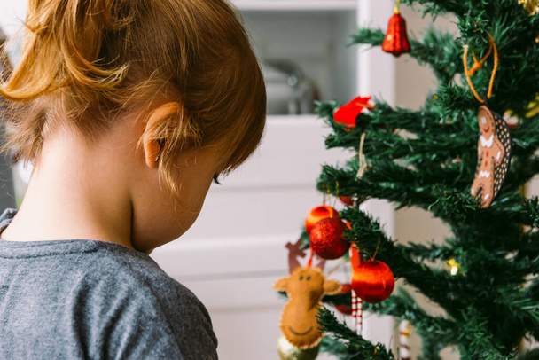 Little blond girl with her back turned decorating the christmas tree at home - Foto, Bild