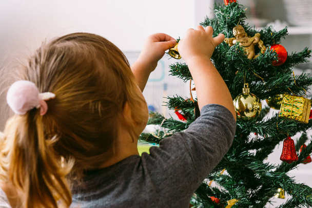 Маленькая блондинка, повернутая спиной, украшает рождественскую елку дома - Фото, изображение