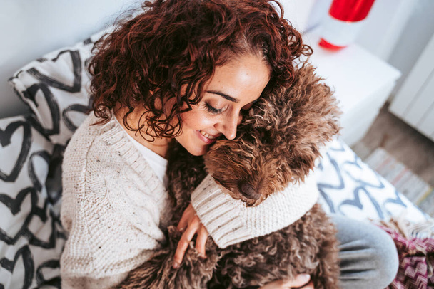 Eine schöne brünette Frau, die sich mit ihrem Hund auf das Bett setzt und ihn umarmt. Sie lächelt und das Haustier ist ruhig. Familienhund-Weihnachtskonzept - Foto, Bild