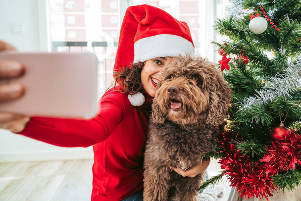 Egy gyönyörű barna nő szórakozik a spanyol vízi kutyájával, miközben fotózza a telefonjával. Télapó sapkát és piros pulóvert visel. Családi kutya karácsonyfa koncepció - Fotó, kép
