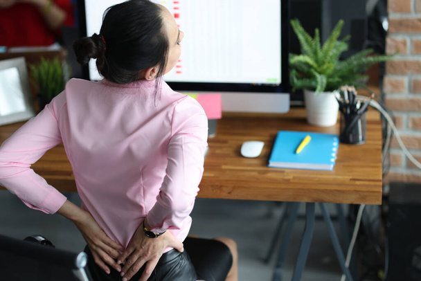 Young woman holding her sore lower back with hands at workplace at table in office - Foto, afbeelding