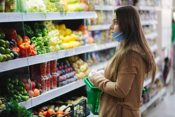 Gelukkige vrouw met een beschermend masker die verse groenten neemt terwijl ze bij de boodschappen staat in de supermarkt. Mooi jong meisje met voedsel mandje kiezen van voedsel door stand met groenten. Kerstaankopen - Foto, afbeelding