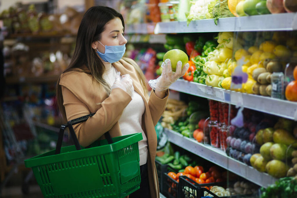 Szczęśliwa kobieta w masce ochronnej biorąc świeże warzywa stojąc przy warzywach w supermarkecie. Piękna młoda dziewczyna z koszykiem żywności wybierając jedzenie na stoisku z owocami. Zakupy świąteczne - Zdjęcie, obraz