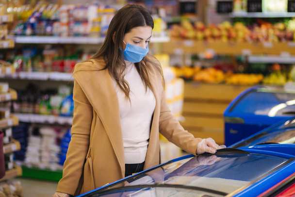 Female shopping during the coronavirus Covid-19 pandemic. A young woman buys semi finished food from refregerator in a supermarket with shopping cart. Woman in facial mask and gloves to prevent - Photo, Image