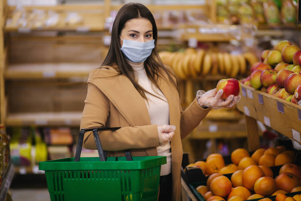 Young woman in protective gloves and face mask hold beautiful fresh apple in hand. Beautiful young girl with food basket choosing food by stand with fruits. Shopping during quarantine. Covid-19 - Photo, image