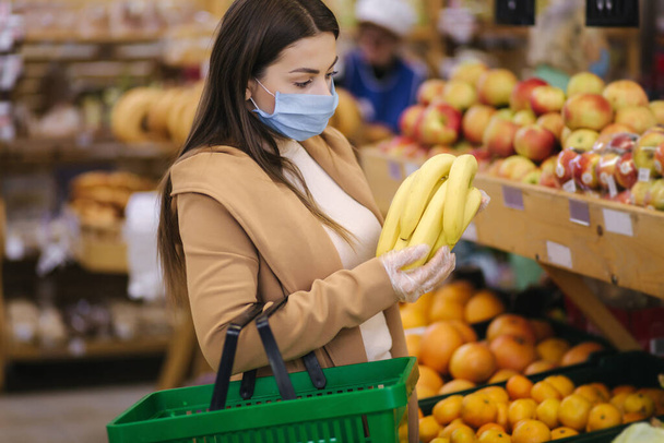 Young woman in protective gloves and face mask hold beautiful fresh bananas in hand. Beautiful young girl with food basket choosing food by stand with fruits. Shopping during quarantine. Covid-19 - Photo, image