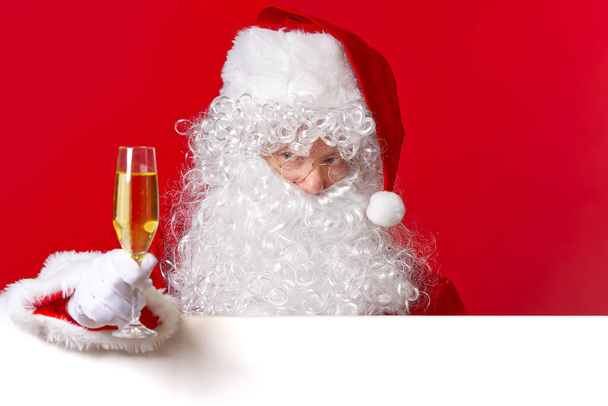 Der Weihnachtsmann hält ein Glas Sekt in der Hand, während er hinter der leeren weißen Tafel steht. Kopierraum. - Foto, Bild