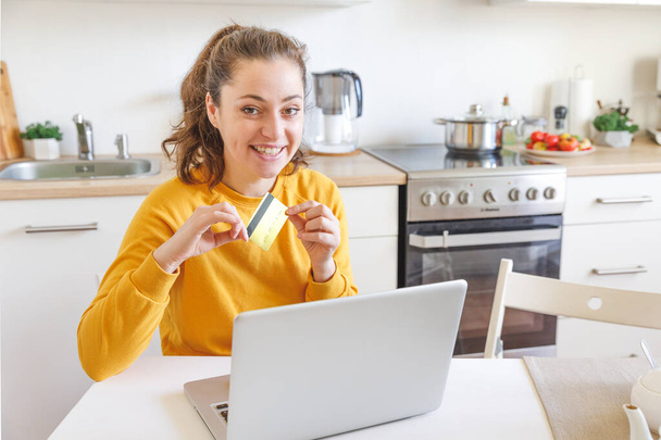 Woman shopping online and paying with gold credit card. Young girl sitting with laptop buying on Internet enter credit card details on kitchen indoor background. Online shopping e-commerce concept - Valokuva, kuva
