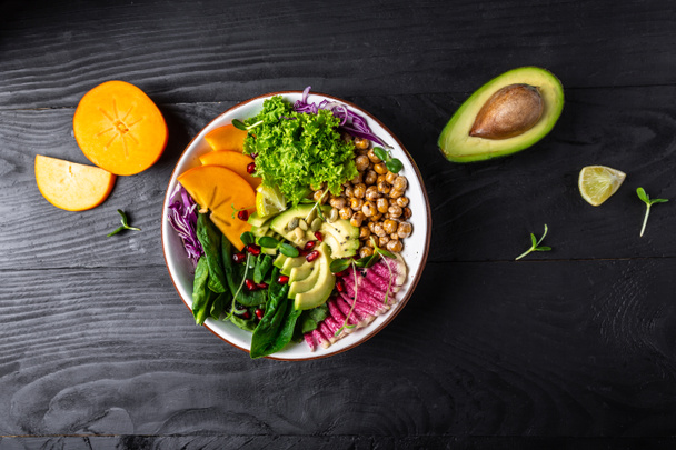 Buddha bowl, healthy and balanced food. roasted chickpeas, avocado, persimmon, spinach, avocado, watermelon radish and seeds top view. - Photo, Image