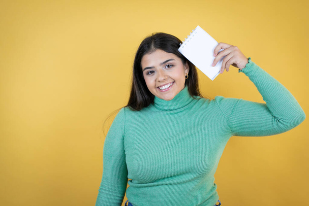 Joven mujer caucásica sobre fondo amarillo aislado sonriendo y mostrando cuaderno en blanco en su mano - Foto, Imagen