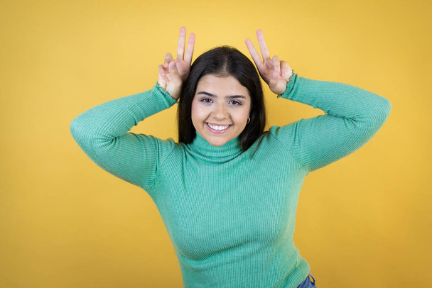 Junge kaukasische Frau auf isoliertem gelben Hintergrund posiert lustig und verrückt mit den Fingern auf dem Kopf als Hasenohren, fröhlich lächelnd - Foto, Bild