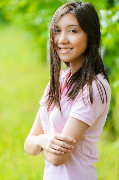 緑色の背景に濃い髪とピンクのブラウスを持つアジアの外観の美しい若い女性夏の自然. - 写真・画像