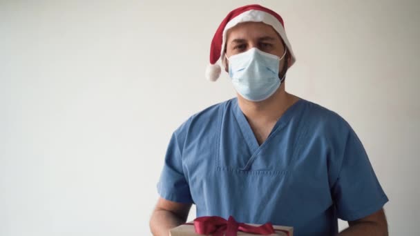 Arzt Kaukasier mit medizinischer Maske und Weihnachtsmann-Hut. Er hält eine Schachtel mit einer roten Schleife in der Hand und hält sie heraus. Konzept eines Geschenks von Ärzten - Filmmaterial, Video