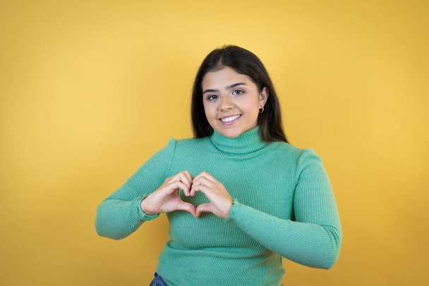 Młoda kaukaska kobieta nad odizolowanym żółtym tle uśmiecha się w miłości pokazując symbol serca i kształt z rąk - Zdjęcie, obraz